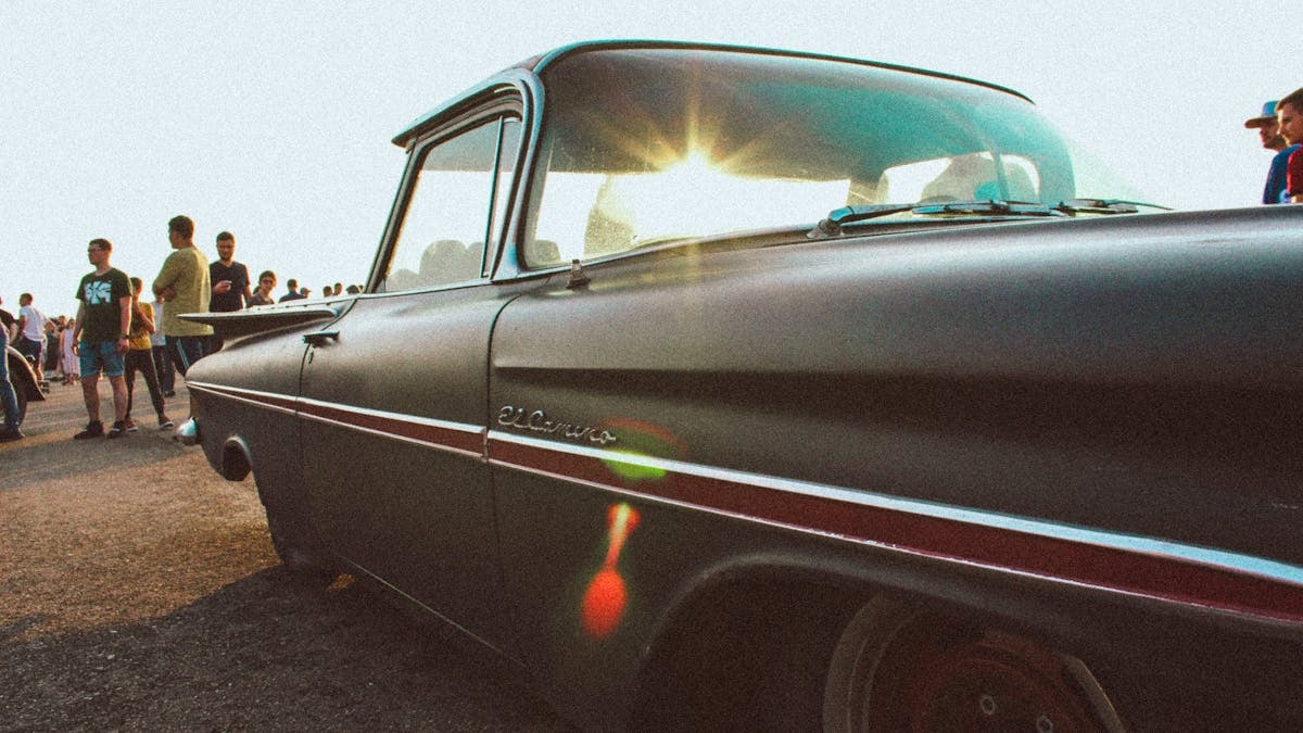Classic Chevrolet El Camino parked under bright sunlight, surrounded by people at an outdoor event.